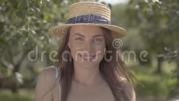 可爱的时尚女青年戴着草帽穿着白色连衣裙看着站在绿色夏日花园里的相机视频的预览图