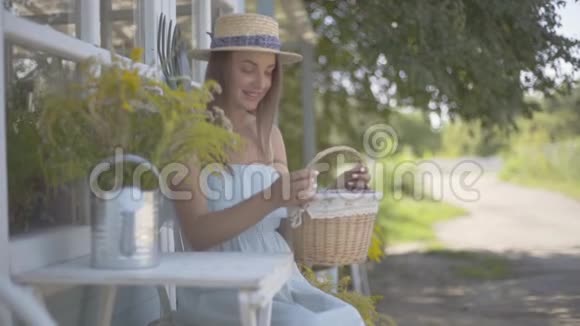 漂亮的年轻女子戴着草帽穿着白色连衣裙把小篮子放在桌子上坐在小桌前视频的预览图