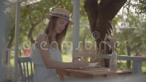 可爱的农村女孩戴着草帽穿着白色连衣裙坐在户外的小木桌上看书视频的预览图