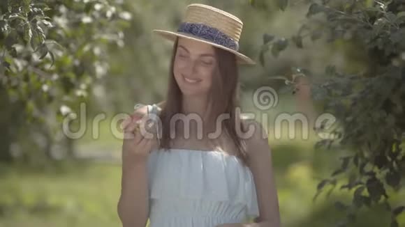 可爱的时尚年轻积极的女人戴着草帽和白色连衣裙看着相机微笑着站着苹果视频的预览图