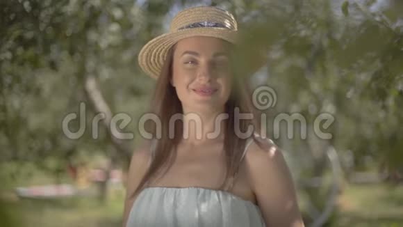 画中漂亮的年轻女子戴着草帽穿着白色长裙看着镜头微笑地站在绿色的夏天视频的预览图