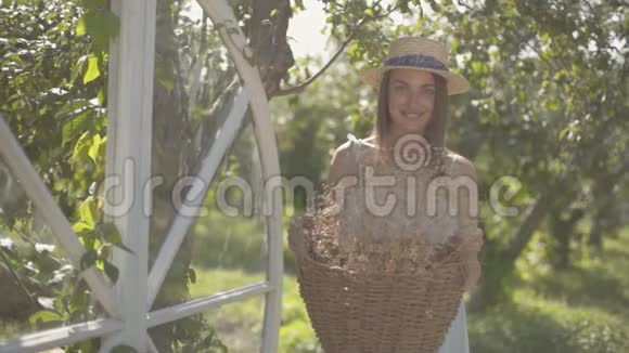可爱的年轻女子戴着草帽拿着柳条篮子拿着草药看着镜头在绿色的夏天微笑着视频的预览图