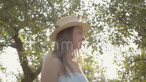 可爱的年轻女子戴着草帽穿着白色的长裙站在绿色的夏日花园里太阳照在女孩身后视频的预览图