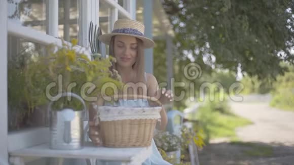 可爱的年轻女人戴着草帽穿着白色连衣裙从小篮子里拿出苹果坐在小村庄前面视频的预览图