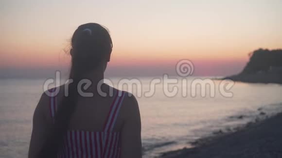 一个年轻的女孩站在海上日落的背景上一个女人背对着镜头站在海滩上视频的预览图
