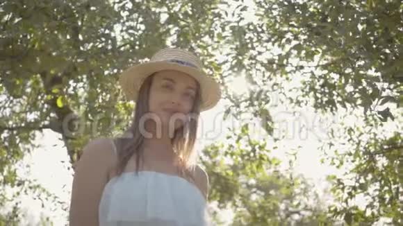 漂亮积极的年轻女子戴着草帽穿着白色长裙看着站在绿色中微笑的镜头视频的预览图