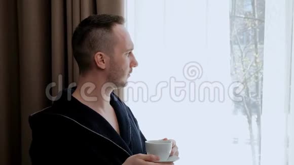 一个穿着浴袍的年轻帅哥站在房间的窗户边从白色的杯子里喝茶或咖啡视频的预览图