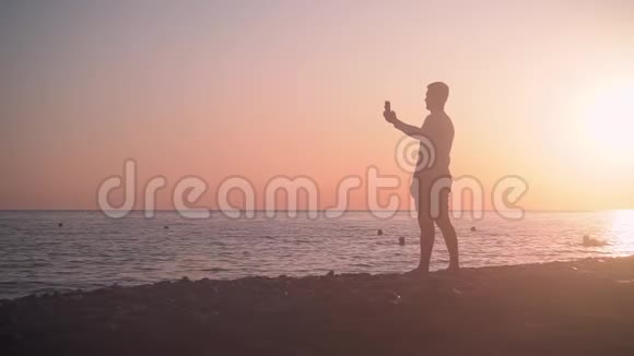 一个年轻人站在海岸上背景是海上日落人在日落背景下在海滩上拍照视频的预览图