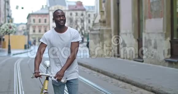 帅气的肌肉发达的非裔美国年轻人戴着耳机环顾四周一边推着时髦的现代自行车视频的预览图