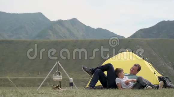 在晴朗的天气里父亲和儿子躺在帐篷前休息在群山的背景下休息视频的预览图