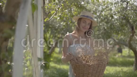 戴着草帽的漂亮年轻女人拿着柳条篮子拿着草药看着镜头在绿色中微笑视频的预览图