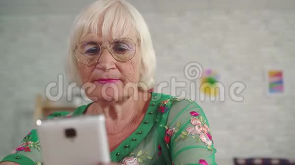 头发灰白的老年妇女使用智能手机坐在靠近客厅的桌子旁视频的预览图