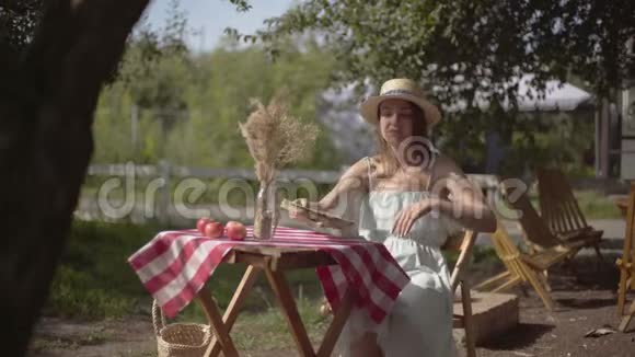 年轻的女孩戴着草帽穿着白色连衣裙端详着看那本带花瓶的小桌子旁的旧书视频的预览图
