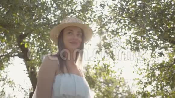 漂亮的年轻女人戴着草帽穿着长长的白色连衣裙站在绿色的夏天花园里太阳照在女孩身后视频的预览图