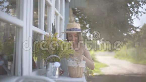 可爱的年轻女子戴着草帽穿着白色连衣裙微笑着坐在浇水罐里嗅着野花视频的预览图