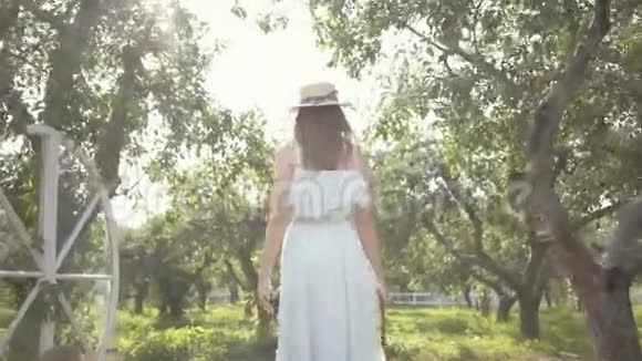 在绿色的夏日花园里美丽的年轻女子戴着草帽穿着长长的白色连衣裙免费视频的预览图
