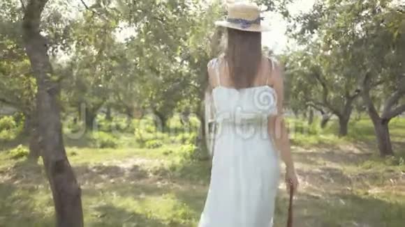 后景可爱的年轻女子戴着草帽和白色长裙走过绿色的夏天花园免费视频的预览图