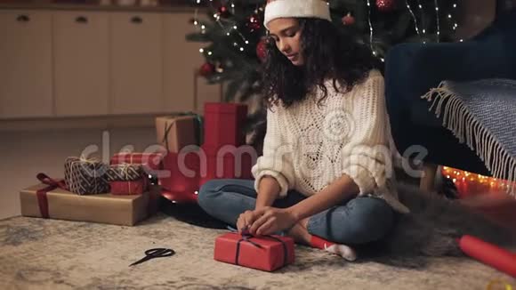 圣诞老人帽子里漂亮的快乐女孩在圣诞树旁准备礼物快乐美丽的女孩在礼物上鞠躬视频的预览图