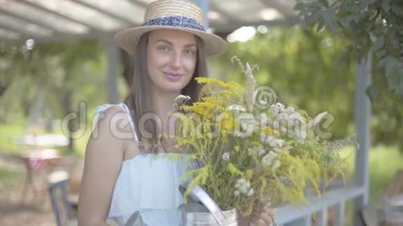 肖像漂亮的年轻女子戴着草帽穿着白色连衣裙看着镜头微笑捧着野花农村视频的预览图