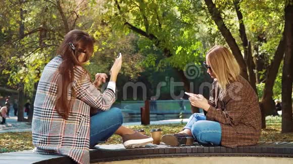在秋季公园时尚女孩面对面坐着使用智能手机视频的预览图