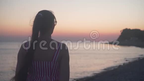 一个年轻的女孩站在海上日落的背景上一个女人背对着镜头站在海滩上视频的预览图