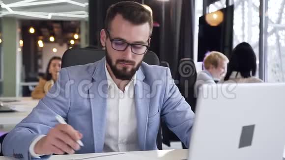 年轻有魅力的商人带着整齐的胡须和眼镜坐在他的工作场所表现得很好视频的预览图