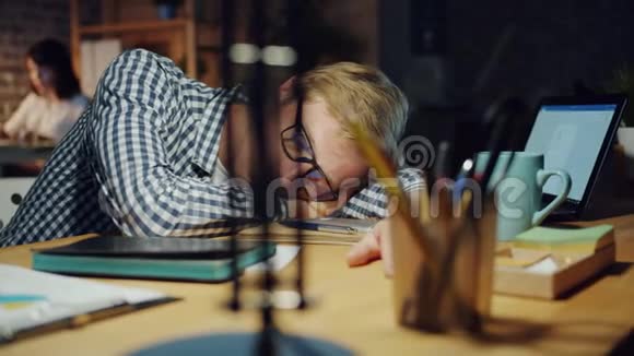 疲倦的商人在办公室放松晚上睡在黑暗的房间里的桌子上视频的预览图