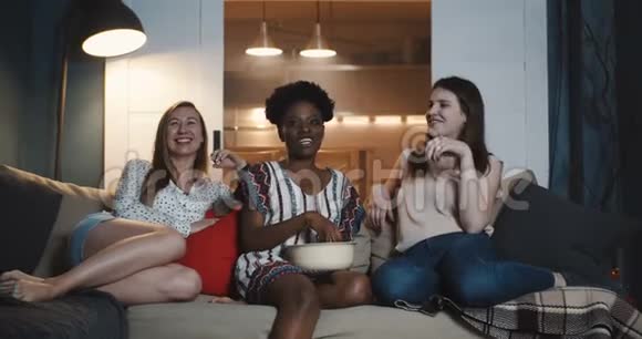 快乐年轻的多民族女性朋友坐在一起看电影微笑着一起吃零食慢动作视频的预览图