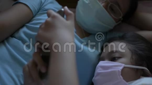 母亲和女儿戴着防护面罩一起躺在床上女儿使用手机视频的预览图
