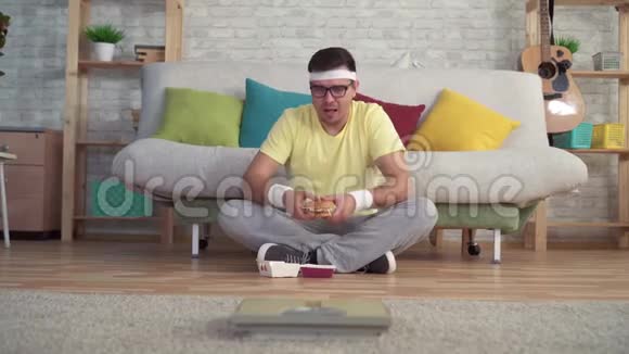 穿着运动服的滑稽男子坐在地板上看着浴室的秤吃着汉堡视频的预览图