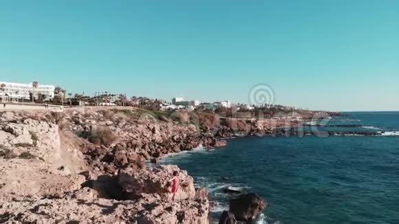 穿着红色衣服的女孩站在岩石悬崖上双臂交叉白色强烈的海浪撞击海岸形成白色泡沫和飞溅视频的预览图