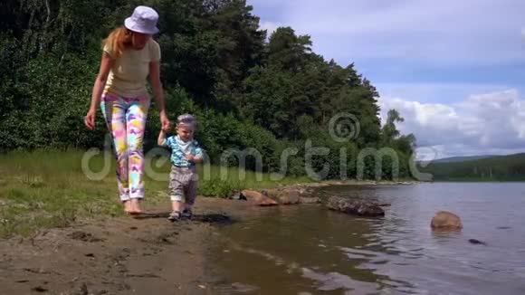 一个夏天妈妈和她的小儿子在森林和蓝天的背景下沿着森林湖的岸边散步视频的预览图