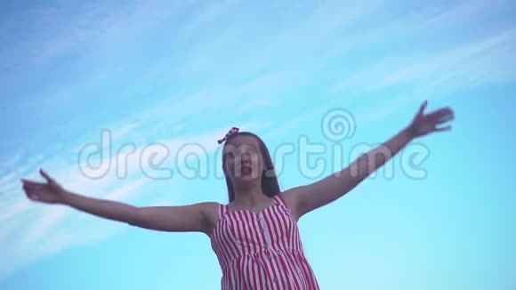 穿着条纹红白连衣裙的年轻女孩一个女孩站在蓝天上举起双手象征着自由视频的预览图