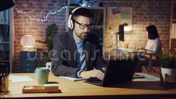 戴着耳机的家伙在办公室里享受音乐晚上忙着用笔记本电脑工作视频的预览图