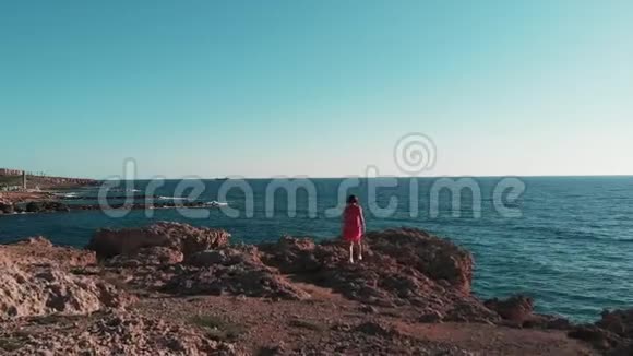 美丽的女孩走在大石头上高加索女人站在岩石海滩上穿红色衣服的女士举起手臂向两边女孩在肖尔视频的预览图