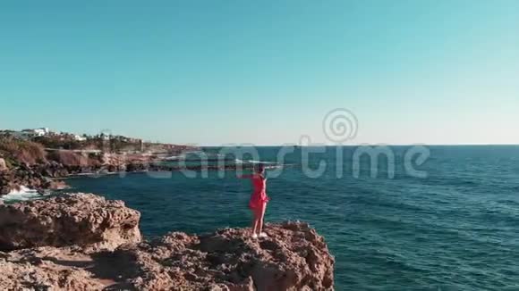 美丽的女孩穿着红色的裙子站在岩石悬崖上年轻女孩站在岩石海滩上双手分开穿短裙子的女孩视频的预览图