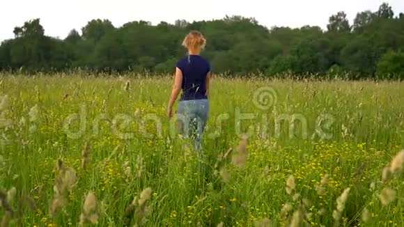 夕阳西下一个穿着蓝色牛仔裤和蓝色t恤的苗条女人在高高的草地上慢慢地穿过田野观点视频的预览图