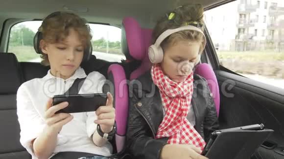 男孩和女孩带着耳机在汽车上玩平板电脑和智能手机孩子们在汽车上使用设备兄弟和兄弟视频的预览图