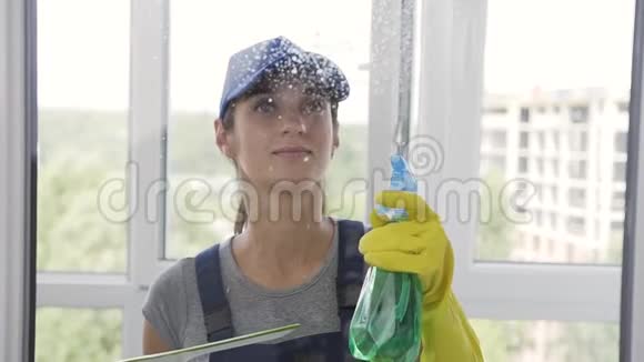 穿着蓝色制服和橡胶手套的开朗女人用喷雾和特殊工具清洁窗户视频的预览图