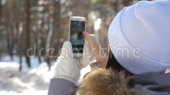 无法辨认的女孩戴着白色帽子在她的智能手机上拍下了美丽的雪林女人用手机视频的预览图