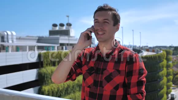 一个穿着格子衬衫的年轻人微笑着在一栋建筑的屋顶上用手机说话背景是视频的预览图