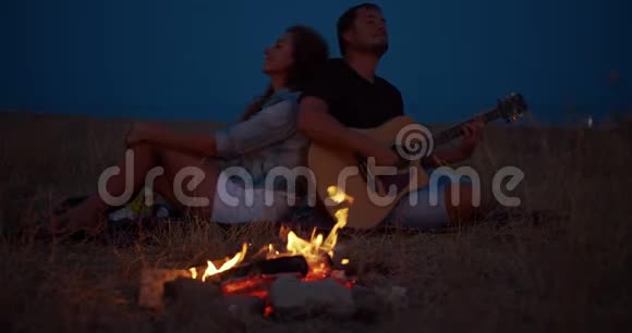 年轻夫妇坐在篝火旁男人弹吉他浪漫的音乐和幸福的夫妻关系浪漫夫妇视频的预览图