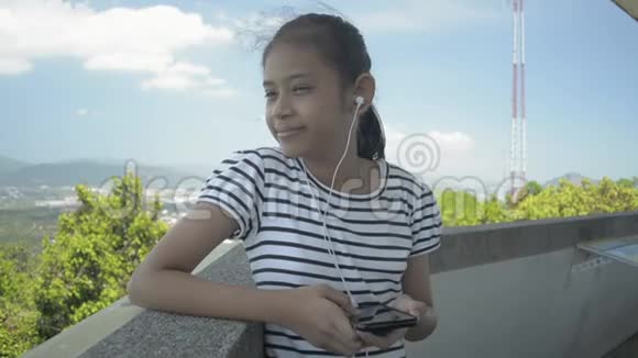 年轻的亚洲女孩愉快地从手机里听音乐站在风景优美的观景塔上视频的预览图