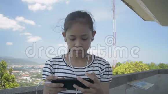 夏天亚洲小女孩用耳机听手机音乐悠闲地在观景塔上散步视频的预览图