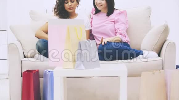 两个快乐的女朋友坐在沙发上在家里用笔记本电脑上网购物模糊的袋子在最前面视频的预览图