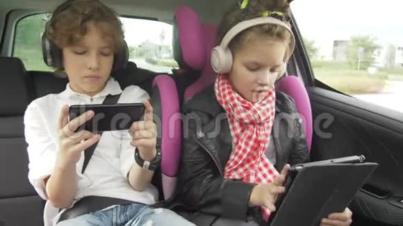 男孩和女孩带着耳机在汽车上玩平板电脑和智能手机孩子们在汽车上使用设备兄弟和兄弟视频的预览图