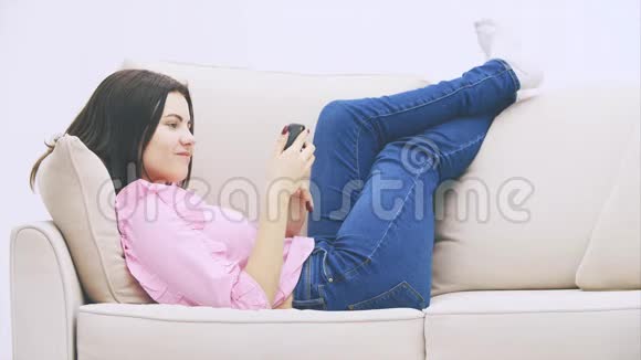 可爱的白种人女孩躺在沙发上把腿放在沙发后面用电话短信视频的预览图