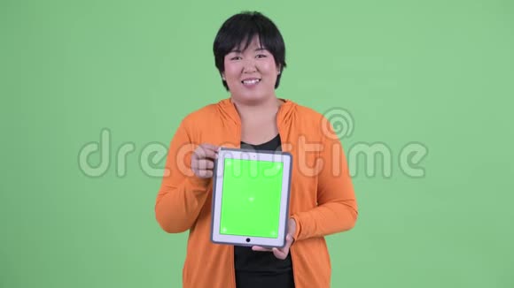快乐年轻体重超重的亚洲女人一边说话一边展示数字平板电脑准备健身房视频的预览图