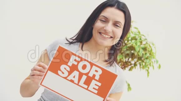 微笑着的年轻美丽的女人拿着一个盘子上面写着出售的文字提供出售的房子并搬家视频的预览图