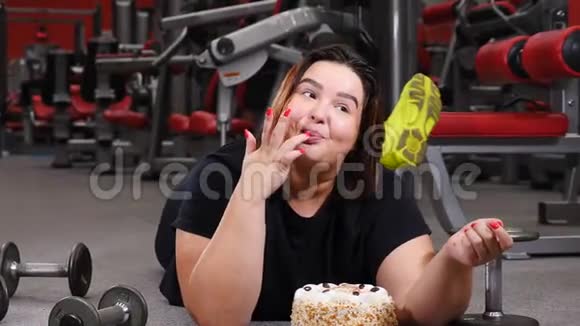 年轻漂亮的肥胖女人躺在健身房地板上用手指蘸着美味的蛋糕意志力概念真实性视频的预览图